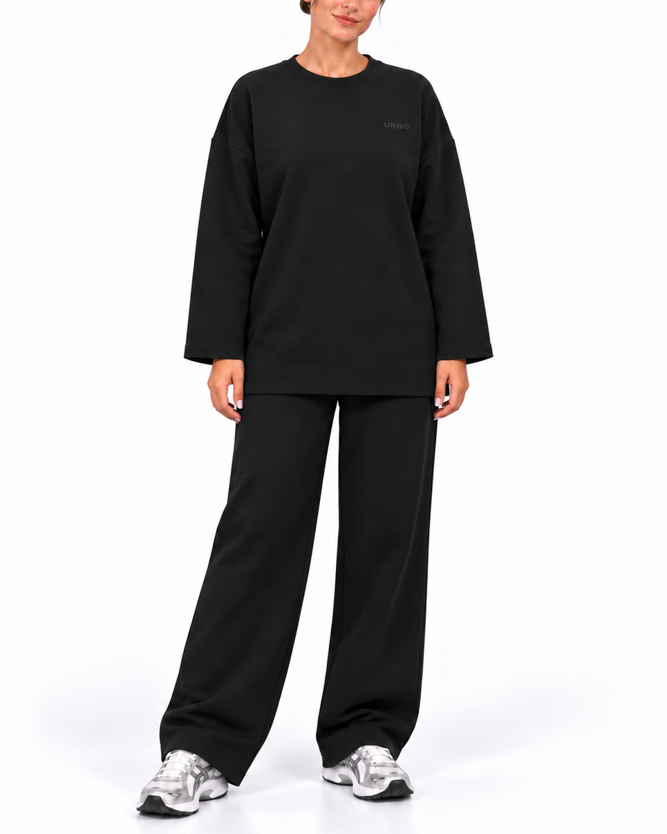A model wearing a black long-sleeve crewneck sweatshirt and matching black wide-leg sweatpants, paired with silver sneakers, against a plain white studio background.