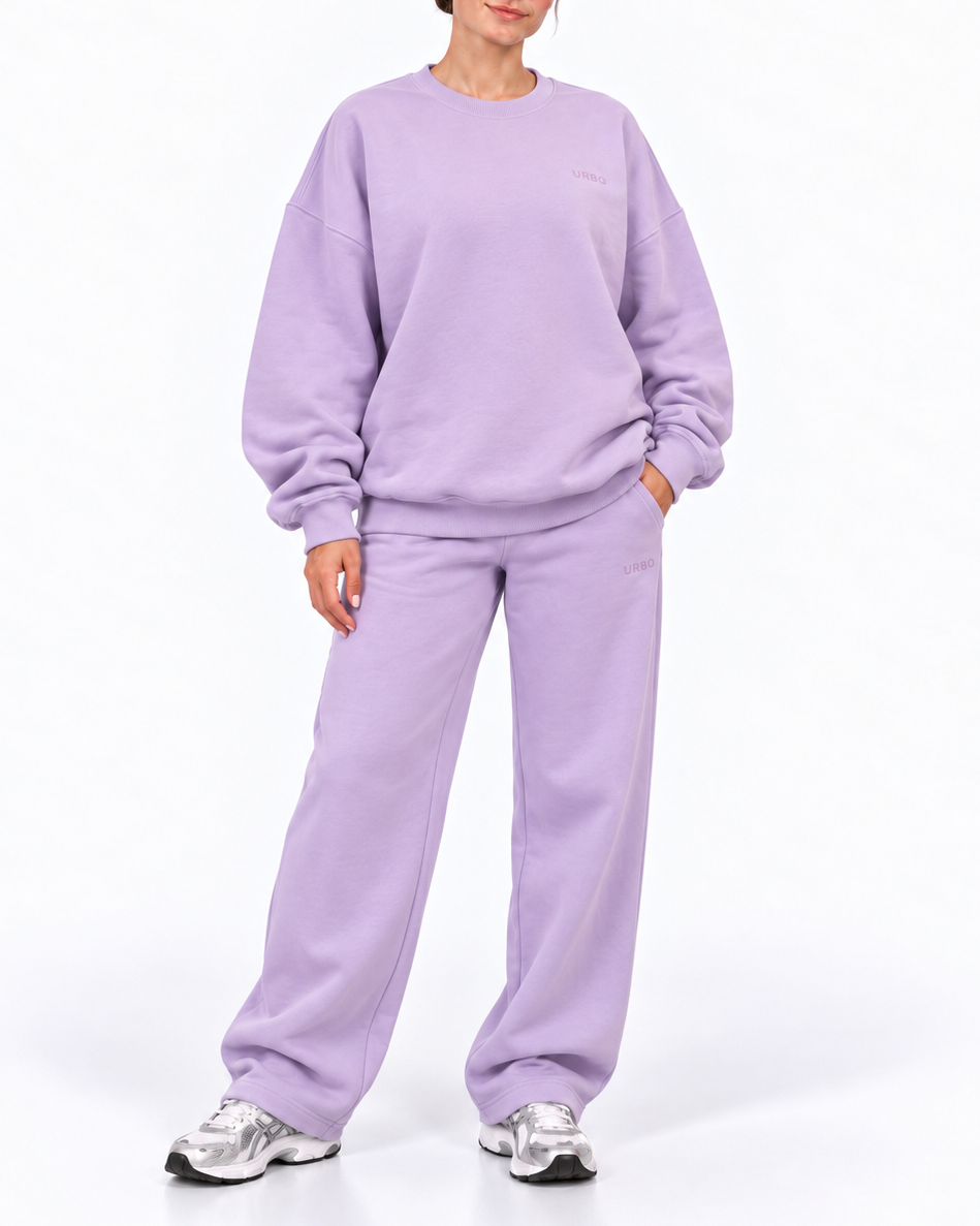 A woman modeling a matching lavender sweatshirt and sweatpants set with silver sneakers, standing against a plain white studio background.