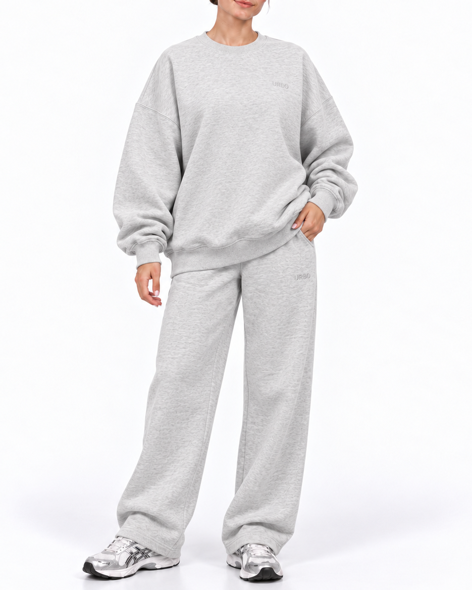 A woman modeling a light gray heather crewneck sweatshirt and matching sweatpants set, paired with gray and white sneakers, against a plain white studio background.