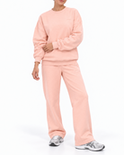 A woman modeling a matching two-piece lounge set in a soft light pink color, consisting of a long-sleeve sweatshirt and wide-leg sweatpants, paired with silver sneakers on a white studio background.
