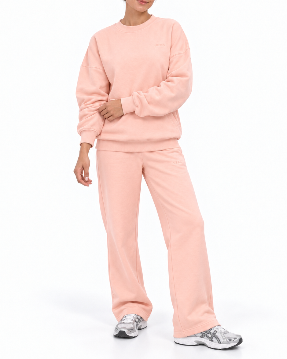 A woman modeling a matching two-piece lounge set in a soft light pink color, consisting of a long-sleeve sweatshirt and wide-leg sweatpants, paired with silver sneakers on a white studio background.