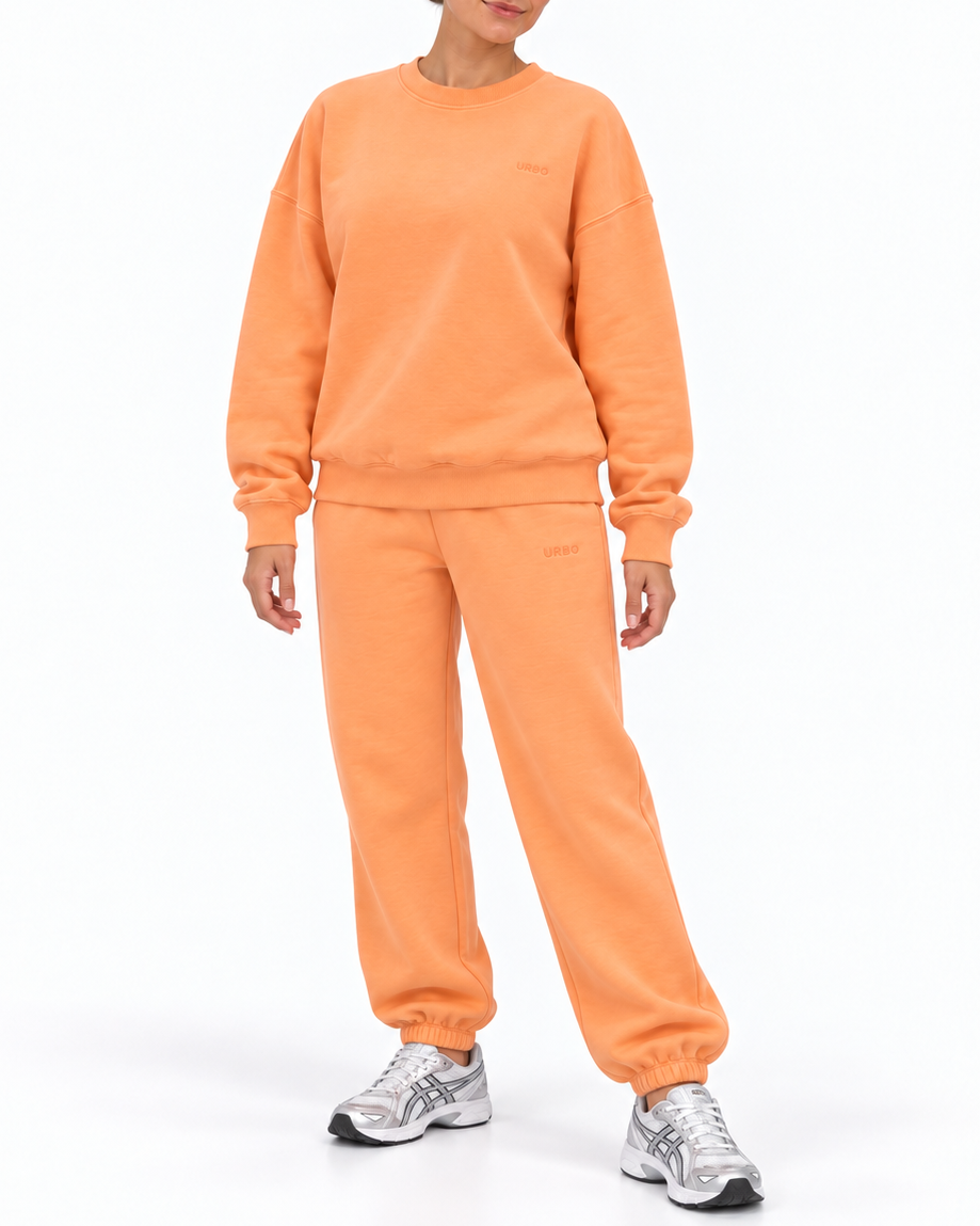 A woman modeling a matching two-piece sweatshirt and jogger pants set in a washed orange color, paired with silver sneakers, against a plain white studio background.