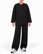 A model wearing a black long-sleeve crewneck sweatshirt and matching black wide-leg sweatpants, paired with gray and white sneakers, against a plain white studio background.