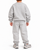 A child viewed from the back wearing a light gray heather sweatshirt and matching sweatpants, standing against a plain white studio background.