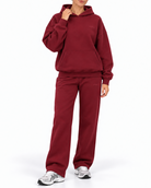A woman modeling a matching burgundy fleece tracksuit, featuring a pullover hoodie and wide-leg sweatpants, paired with gray and white sneakers, against a plain white studio background.