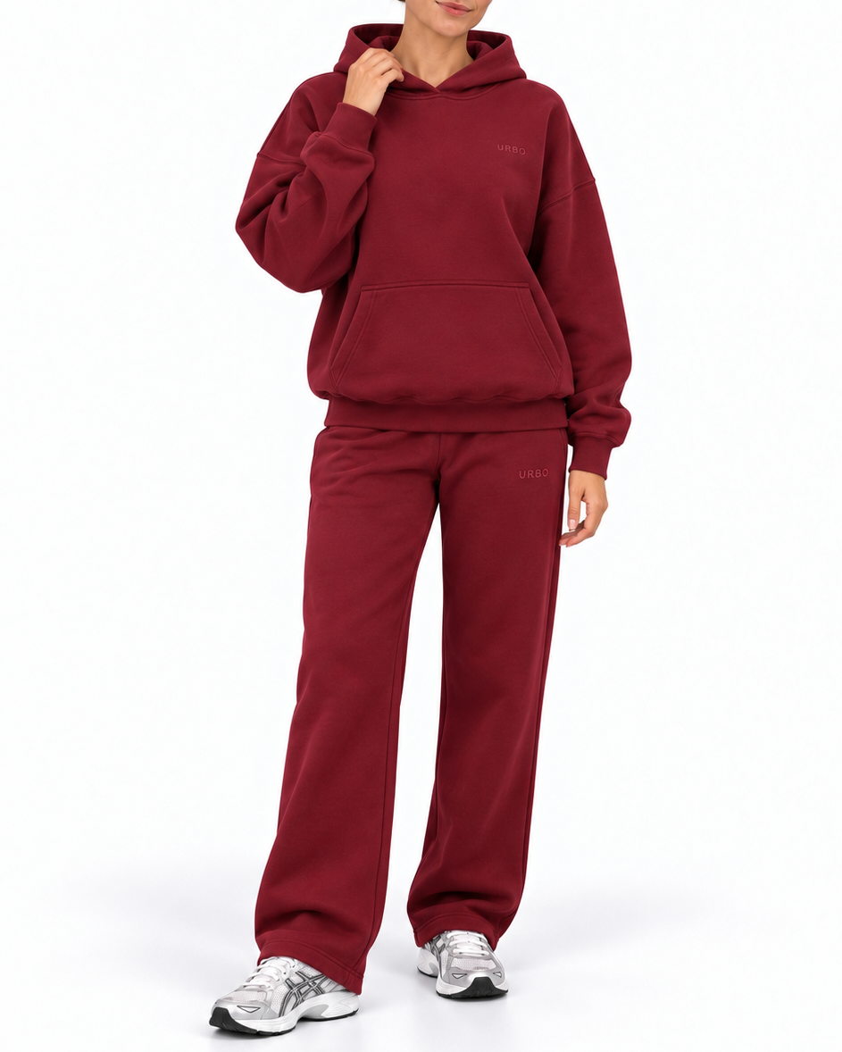 A woman modeling a matching burgundy fleece tracksuit, featuring a pullover hoodie and wide-leg sweatpants, paired with gray and white sneakers, against a plain white studio background.