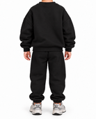 A child viewed from the back wearing a black crewneck sweatshirt and matching black sweatpants, paired with gray and white sneakers, against a plain white background.