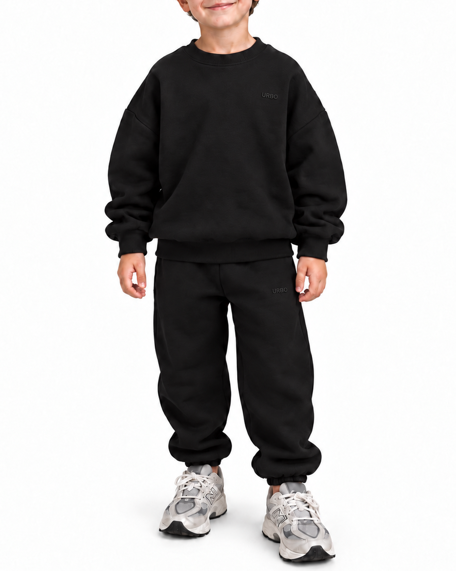 A young boy wearing a black crewneck sweatshirt and matching black sweatpants, paired with gray and white sneakers, standing against a plain white background.