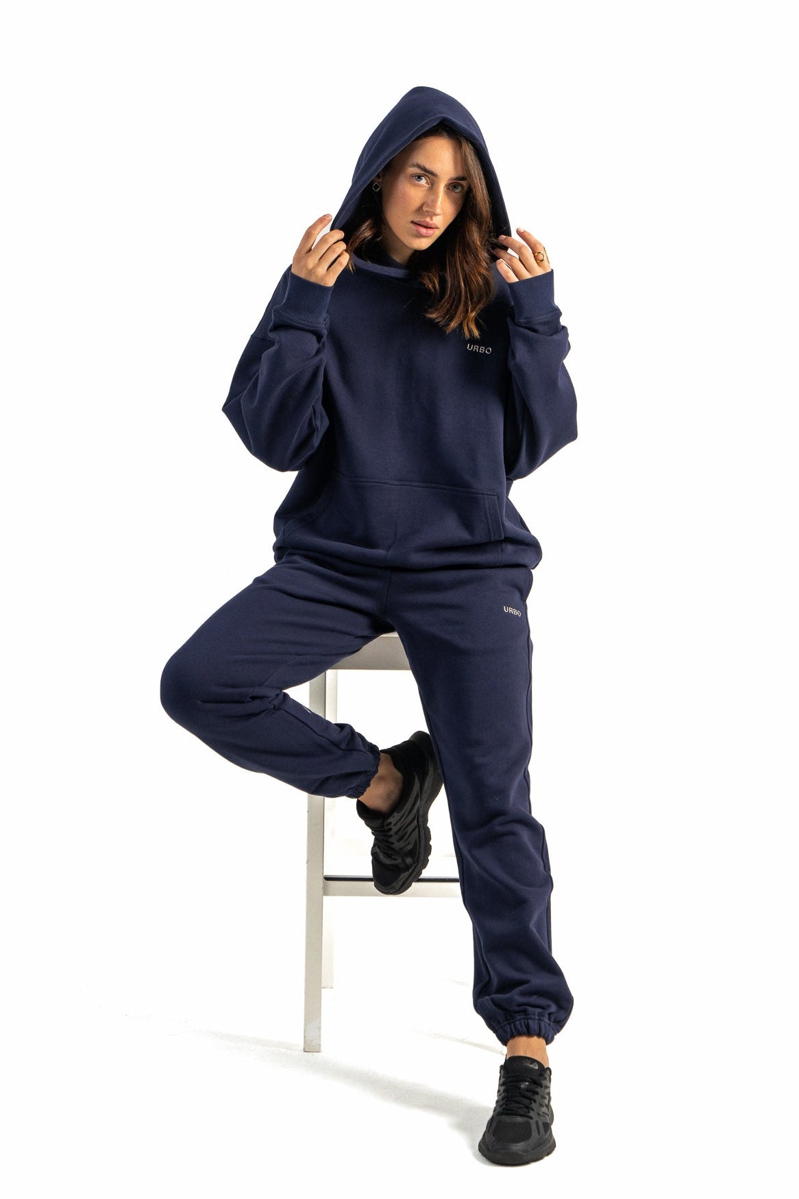 Person wearing a navy blue hoodie and pants sitting on a white stool against a white background