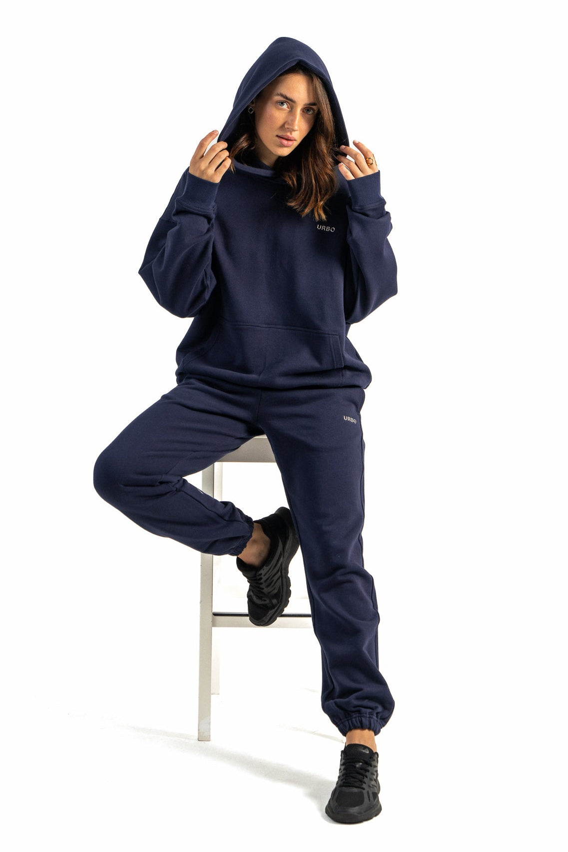 Person wearing a navy blue hoodie and pants sitting on a white stool against a white background