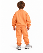 A child viewed from the back wearing a matching orange sweatshirt and sweatpants set with gray sneakers, photographed against a plain white studio background.