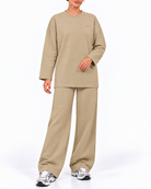 A woman modeling a matching beige lounge set consisting of a long-sleeve crewneck sweatshirt and wide-leg sweatpants, paired with silver sneakers, against a plain white studio background.