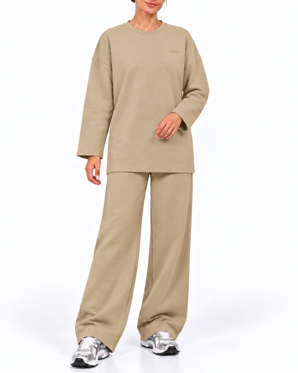 A woman modeling a matching beige lounge set consisting of a long-sleeve crewneck sweatshirt and wide-leg sweatpants, paired with silver sneakers, against a plain white studio background.