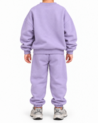 A child viewed from the back wearing a matching light purple sweatshirt and jogger pants set, paired with gray sneakers, against a plain white background.
